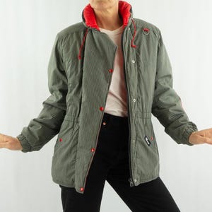 Op de afbeelding: Een groen en wit gestreepte jas met een rode kraag en rode accenten op de knopen en capuchonkoorden. De jas is dichtgeknoopt en heeft een taille met trekkoord. De jas wordt over een lichtgekleurd shirt gedragen.