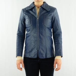 K&ouml;nnte beinhalten: Eine marineblaue Vintage-Lederjacke mit Kragen und durchgehendem Rei&szlig;verschluss. Die Jacke hat zwei Brusttaschen und eine taillierte Passform. Die &Auml;rmel sind lang, und die Jacke wird &uuml;ber einem cremefarbenen Pullover getragen.