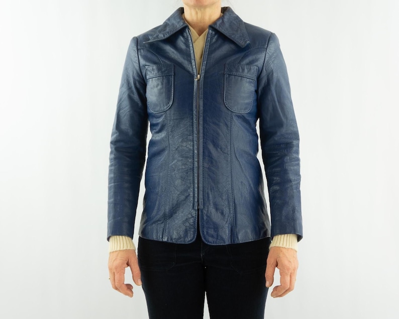 K&ouml;nnte beinhalten: Eine marineblaue Vintage-Lederjacke mit Kragen und durchgehendem Rei&szlig;verschluss. Die Jacke hat zwei Brusttaschen und eine taillierte Passform. Die &Auml;rmel sind lang, und die Jacke wird &uuml;ber einem cremefarbenen Pullover getragen.