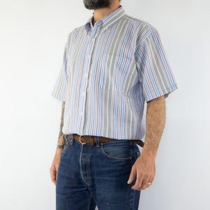 Peut inclure: Une chemise à manches courtes à rayures, avec des rayures bleues, rouges et grises. La chemise a une poche poitrine et une ceinture en cuir marron.