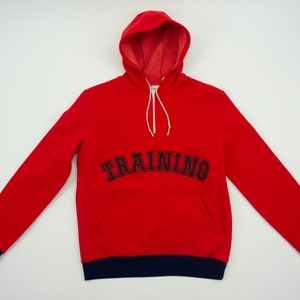 K&ouml;nnte beinhalten: Roter Kapuzenpullover mit dunkelblauem Saum und dem Wort "TRAINING" in dunkelblau auf der Vorderseite gestickt.