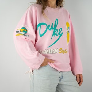 Puede incluir: Sudadera rosa con un estampado gráfico de la palabra "Duke" en turquesa y amarillo, con el texto "Hawaiian Style" debajo. La sudadera tiene un cordón en la parte inferior.
