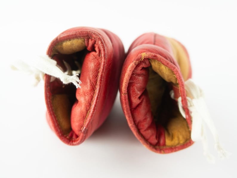 May include: Pair of vintage red leather boxing gloves with white laces. The gloves have a worn appearance, with visible stitching and a yellowed interior. The gloves are set against a white background.