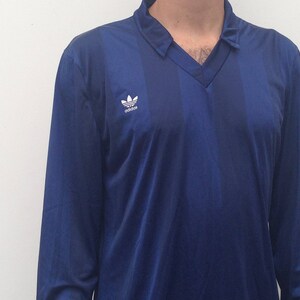 K&ouml;nnte beinhalten: Ein lang&auml;rmliges, marineblaues Hemd mit einem wei&szlig;en Adidas-Logo auf der linken Brust. Das Hemd hat einen V-Ausschnitt.