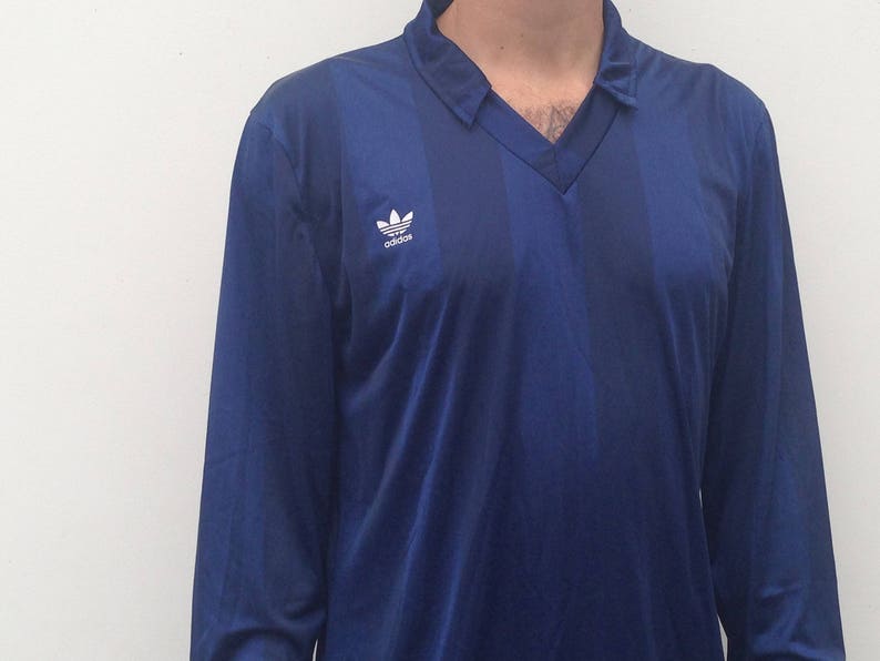 K&ouml;nnte beinhalten: Ein lang&auml;rmliges, marineblaues Hemd mit einem wei&szlig;en Adidas-Logo auf der linken Brust. Das Hemd hat einen V-Ausschnitt.