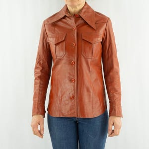 May include: A vintage, rust-colored leather shirt with a pointed collar and two chest pockets. The long-sleeved shirt has button closures down the front and on the cuffs. The model is wearing blue jeans.
