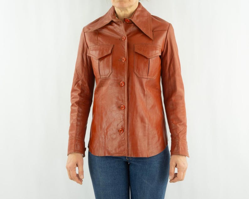 May include: A vintage, rust-colored leather shirt with a pointed collar and two chest pockets. The long-sleeved shirt has button closures down the front and on the cuffs. The model is wearing blue jeans.