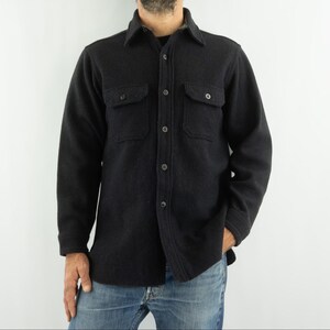 May include: A dark gray button-down shirt with two chest pockets. The long-sleeved shirt is made of a thick, textured fabric and features a classic collar. The shirt is worn with blue jeans.