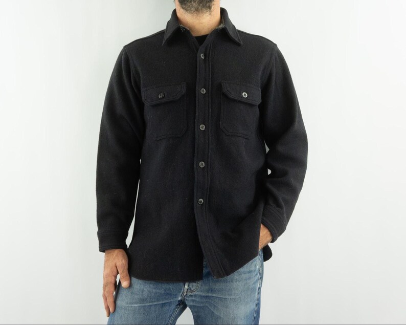 May include: A dark gray button-down shirt with two chest pockets. The long-sleeved shirt is made of a thick, textured fabric and features a classic collar. The shirt is worn with blue jeans.