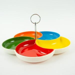 Peut inclure: Un plat de service à sections multiples composé de cinq bols individuels disposés en forme de fleur. Chaque bol est d'une couleur différente : rouge, orange, jaune, vert et bleu. L'extérieur est blanc, avec une poignée centrale en métal argenté.