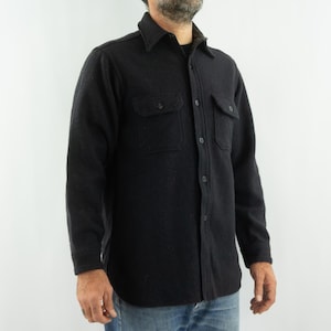 May include: A dark gray button-down shirt with two chest pockets. The shirt has a collared neckline and is made of a textured fabric. The sleeves are long, and the shirt is buttoned up. The shirt is worn over a black undershirt.