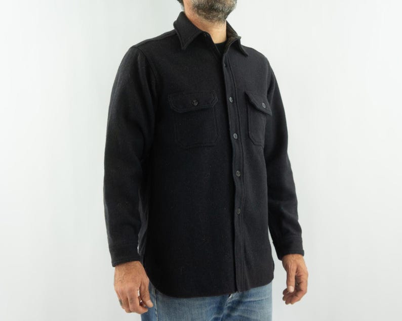 May include: A dark gray button-down shirt with two chest pockets. The shirt has a collared neckline and is made of a textured fabric. The sleeves are long, and the shirt is buttoned up. The shirt is worn over a black undershirt.