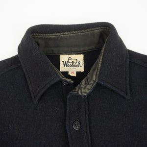 May include: Close-up of a black wool shirt with a collar and button closure. The shirt features a leather-lined collar and a label that reads "Woolrich EST 1830". The shirt is a medium size.