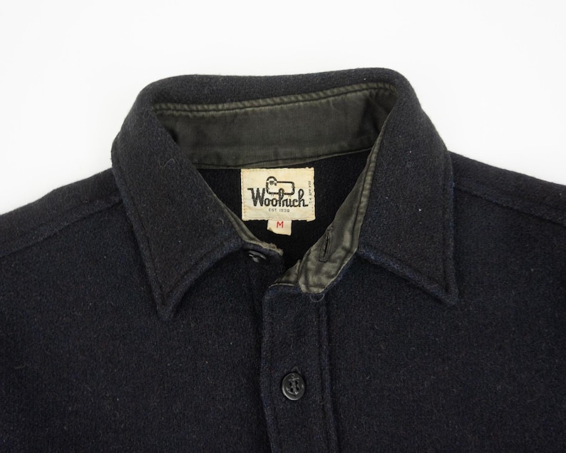 May include: Close-up of a black wool shirt with a collar and button closure. The shirt features a leather-lined collar and a label that reads "Woolrich EST 1830". The shirt is a medium size.