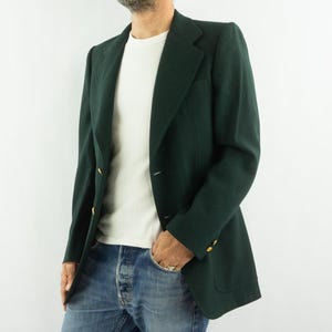 Peut inclure: Un blazer vert foncé avec des boutons dorés. Le blazer a un design classique avec un revers cranté et une poche sur le côté gauche. La personne porte un t-shirt blanc et un jean bleu.