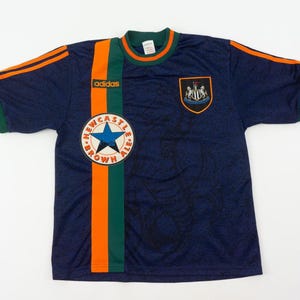 May include: Navy blue soccer jersey with orange and green accents. Features the Adidas logo, a Newcastle Brown Ale patch, and a team crest. The jersey has short sleeves and a round neckline.