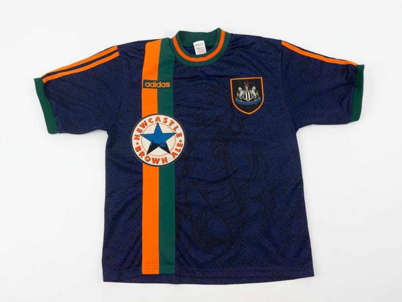 May include: Navy blue soccer jersey with orange and green accents. Features the Adidas logo, a Newcastle Brown Ale patch, and a team crest. The jersey has short sleeves and a round neckline.