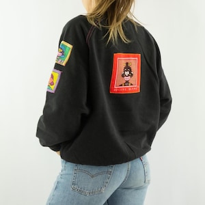 Peut inclure: Sweat-shirt noir avec des écussons colorés sur les manches et le dos. Les écussons présentent des illustrations de type dessin animé avec du texte. Le sweat-shirt est associé à un jean bleu. Le patch arrière porte le texte "POIVRE BLANC".