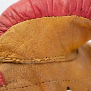 May include: Close-up of a vintage boxing glove. The glove features a red leather top and a tan leather body. The stitching is visible, and the leather shows signs of wear, suggesting it's an antique or well-used item.