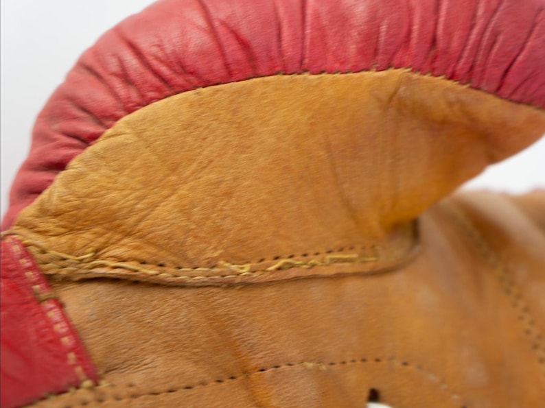 May include: Close-up of a vintage boxing glove. The glove features a red leather top and a tan leather body. The stitching is visible, and the leather shows signs of wear, suggesting it's an antique or well-used item.