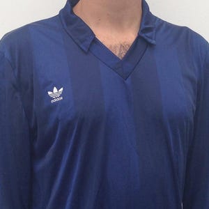 K&ouml;nnte beinhalten: Ein lang&auml;rmeliges Hemd in Dunkelblau mit einem dezenten vertikalen Streifenmuster. Das Hemd hat einen V-Ausschnitt und das wei&szlig;e Adidas-Trefoil-Logo ist auf der rechten Brustseite aufgedruckt. Der Stoff weist einen leichten Glanz auf.