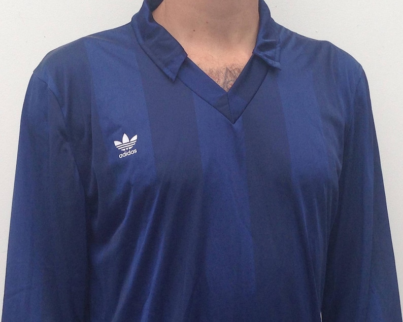 K&ouml;nnte beinhalten: Ein lang&auml;rmeliges Hemd in Dunkelblau mit einem dezenten vertikalen Streifenmuster. Das Hemd hat einen V-Ausschnitt und das wei&szlig;e Adidas-Trefoil-Logo ist auf der rechten Brustseite aufgedruckt. Der Stoff weist einen leichten Glanz auf.