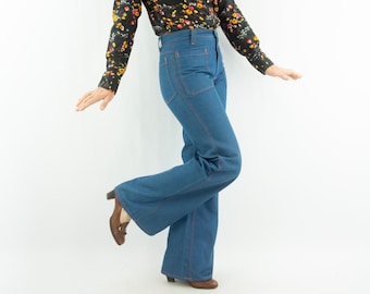 Puede incluir: Vaqueros acampanados de mezclilla azul con costuras rojas. Los vaqueros de cintura alta tienen una pernera ancha. La persona lleva una blusa negra con estampado floral y tacones marrones. Estilo vintage.