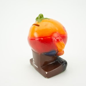 May include: A ceramic piggy bank shaped like a cartoon orange with a green stem and wearing brown pants. The piggy bank has a slot on the back for coins and the words "Save All" are printed on the base.