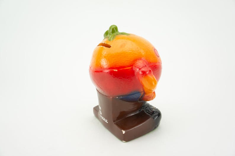 May include: A ceramic piggy bank shaped like a cartoon orange with a green stem and wearing brown pants. The piggy bank has a slot on the back for coins and the words "Save All" are printed on the base.