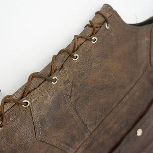 May include: Close-up of a brown leather vest with a distressed finish. The vest features a lace-up closure on the side and a small patch with a graphic. The leather has visible wear and tear.