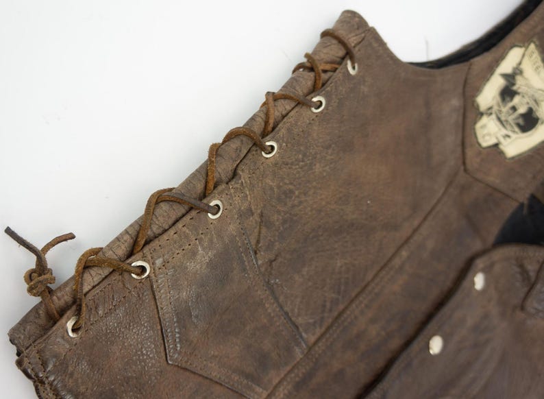 May include: Close-up of a brown leather vest with a distressed finish. The vest features a lace-up closure on the side and a small patch with a graphic. The leather has visible wear and tear.