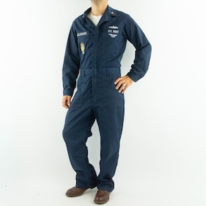 Peut inclure: Combinaison bleu marine de la marine américaine avec des manches longues et un col. La combinaison comporte une étiquette nominative, un emblème de la marine américaine et des poches. La personne porte des chaussures en cuir marron. Uniforme militaire vintage.