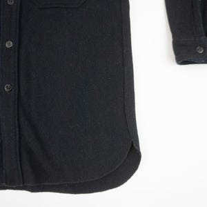 May include: Close-up of a black button-down shirt. The shirt has a classic collar, buttoned cuffs, and two chest pockets. The fabric appears to be a thick, textured material. The buttons are small and dark, blending with the shirt's color.