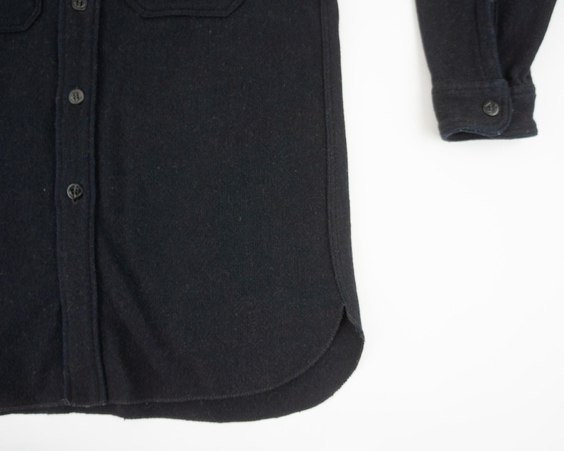 May include: Close-up of a black button-down shirt. The shirt has a classic collar, buttoned cuffs, and two chest pockets. The fabric appears to be a thick, textured material. The buttons are small and dark, blending with the shirt's color.