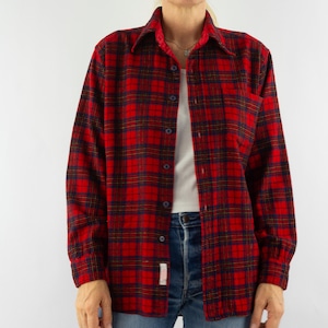 Puede incluir: Una camisa de franela a cuadros rojos y negros con mangas largas y cuello. La camisa tiene una parte delantera abotonada y un bolsillo en el pecho. El patrón de cuadros incluye acentos azul marino y amarillo. La camisa se usa sobre una camiseta blanca y vaqueros azules.