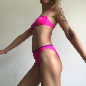 May include: A pink bikini with a purple trim. The top is a bandeau style and the bottom is a high-cut style.