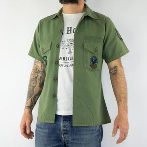 May include: A green short-sleeved button-up shirt with multiple patches. The patches include a green and white patch with a design, a blue and white patch with a bird design, and a green patch with a design. The shirt has a white t-shirt underneath.