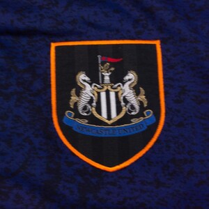 May include: A navy blue fabric with a Newcastle United emblem. The emblem is a shield shape with a black background, orange border, and a blue banner at the bottom with the words "Newcastle United".