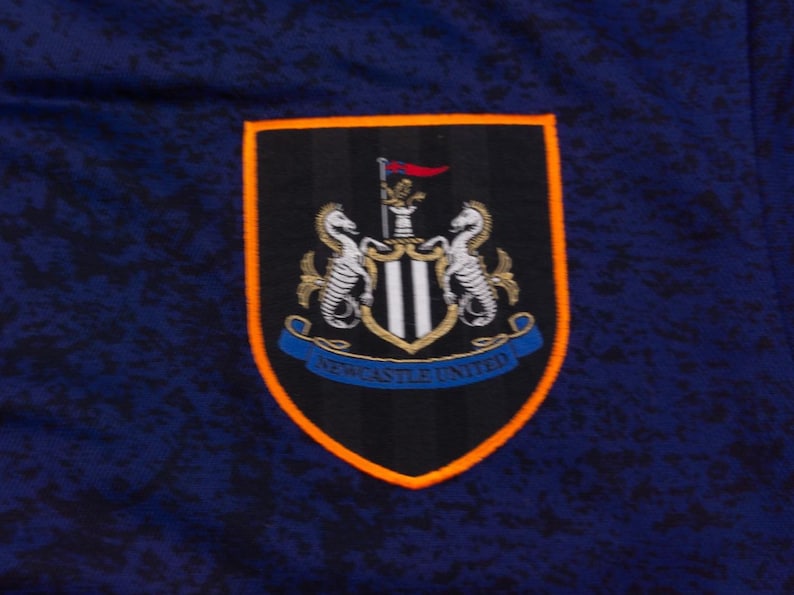 May include: A navy blue fabric with a Newcastle United emblem. The emblem is a shield shape with a black background, orange border, and a blue banner at the bottom with the words "Newcastle United".