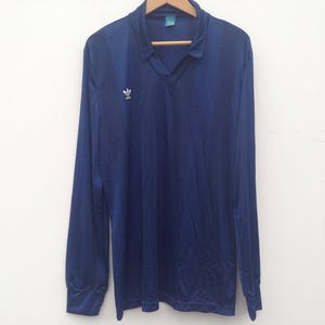 K&ouml;nnte beinhalten: Ein lang&auml;rmeliges, marineblaues Poloshirt mit V-Ausschnitt und einem wei&szlig;en Adidas-Trefoil-Logo auf der rechten Brust. Das Shirt besteht aus einem glatten, leicht reflektierenden Stoff und hat ein kleines Etikett im inneren Kragenbereich.