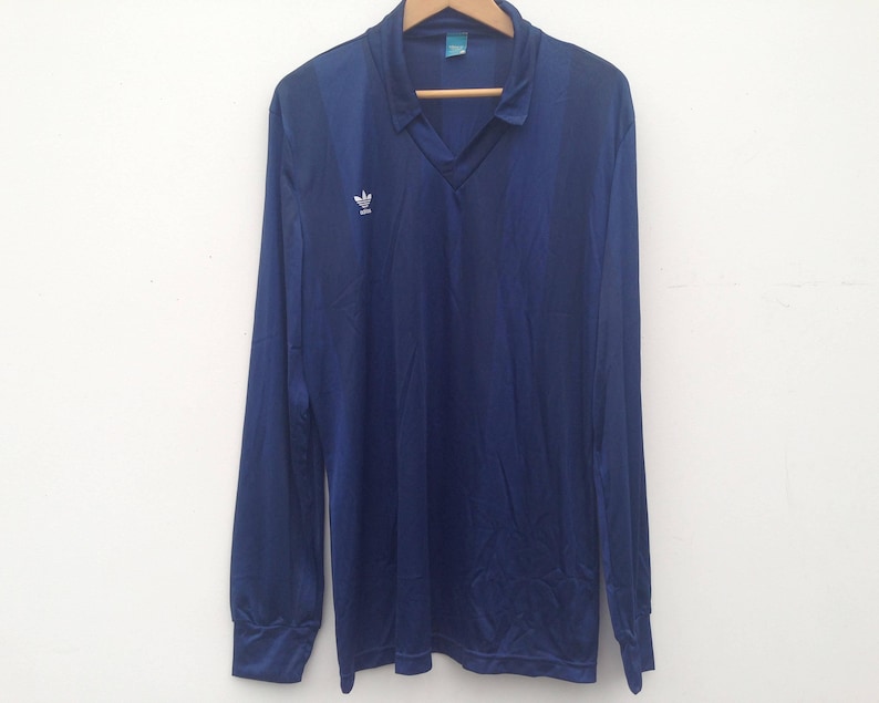 K&ouml;nnte beinhalten: Ein lang&auml;rmeliges, marineblaues Poloshirt mit V-Ausschnitt und einem wei&szlig;en Adidas-Trefoil-Logo auf der rechten Brust. Das Shirt besteht aus einem glatten, leicht reflektierenden Stoff und hat ein kleines Etikett im inneren Kragenbereich.