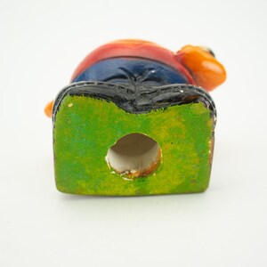 May include: A ceramic figurine of a person with a red, orange, and blue shirt, blue jeans, and black shoes. The figurine has a green base with a hole in the center.