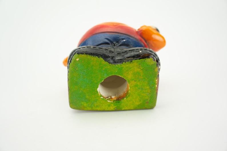 May include: A ceramic figurine of a person with a red, orange, and blue shirt, blue jeans, and black shoes. The figurine has a green base with a hole in the center.