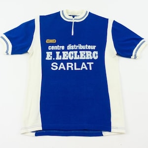 May include: A vintage blue and white cycling jersey with the text "centre distributeur E.LECLERC SARLAT" in white block letters. The jersey has a short zipper at the collar and white trim on the sleeves and collar.
