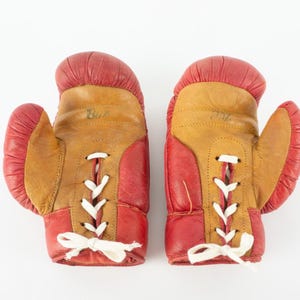 May include: A pair of vintage boxing gloves. The gloves are red and tan leather with white laces. The word "Blue" is written on one glove. The gloves are likely used and show signs of wear.