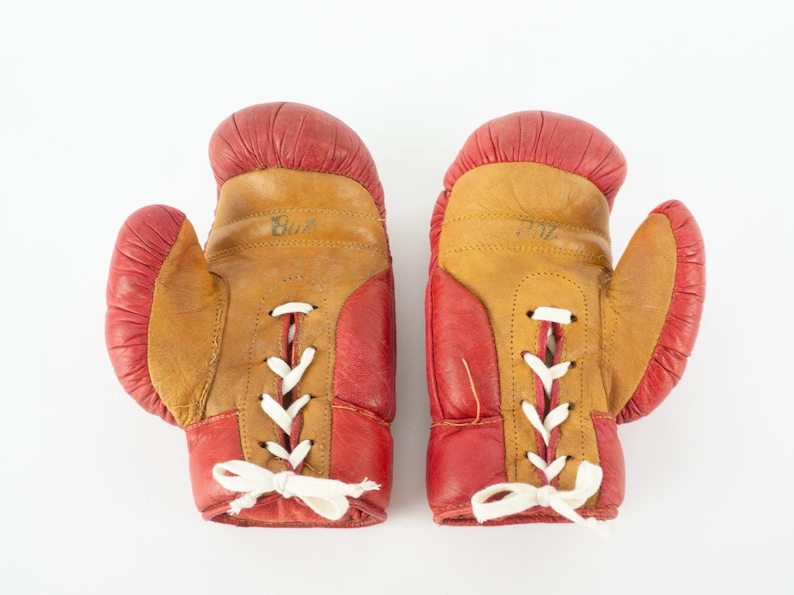 May include: A pair of vintage boxing gloves. The gloves are red and tan leather with white laces. The word "Blue" is written on one glove. The gloves are likely used and show signs of wear.