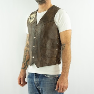 May include: Brown leather vest with a skull patch on the chest. The vest has a button-up front, two front pockets, and lace-up sides. The vest is worn over a white t-shirt and blue jeans.