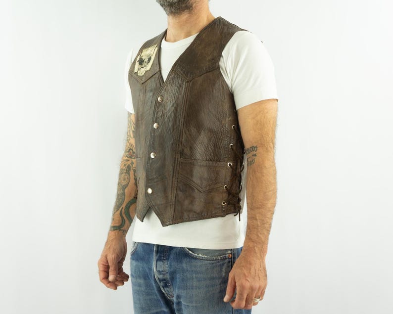 May include: Brown leather vest with a skull patch on the chest. The vest has a button-up front, two front pockets, and lace-up sides. The vest is worn over a white t-shirt and blue jeans.