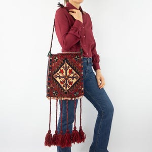 May include: A rectangular shoulder bag crafted from a woven rug featuring a central diamond motif in red, navy blue, and cream. The bag includes a long braided strap and five long braided tassels with thick fringe ends hanging from the bottom edge.