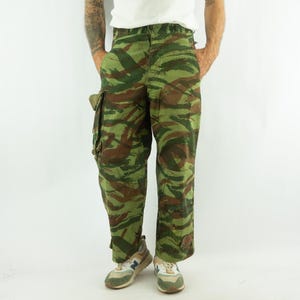 May include: Cargo pants featuring a green, brown, and black tiger stripe camouflage pattern. These trousers include side cargo pockets on the thighs, a button fly closure, and a straight-leg fit, constructed from a durable, woven fabric material.